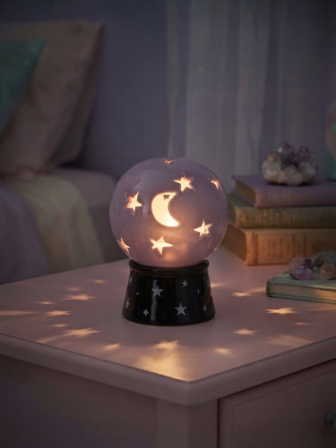 A glowing A pastel lavender crystal ball tea light holder and incense burner with star and moon cutouts sits on a bedside table in a softly lit bedroom. The sphere glows with warm light, casting star-shaped shadows across the surface, while a dark star-patterned base supports it.