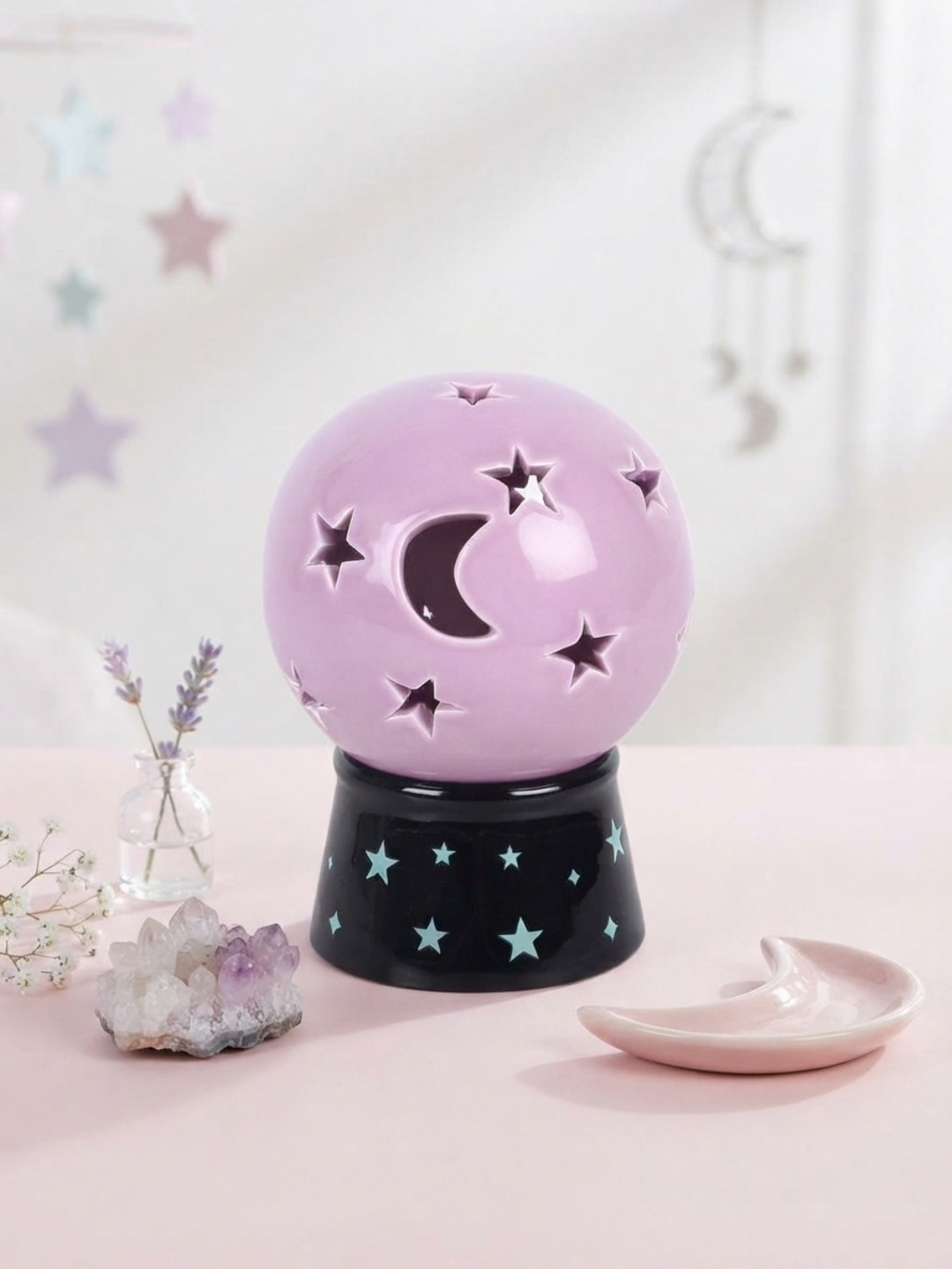 A pastel lavender crystal ball tea light holder and incense burner with star and moon cutouts, sitting on a black base decorated with light blue stars. The scene is styled on a pink surface with a small pink crescent moon dish and crystals in the background.