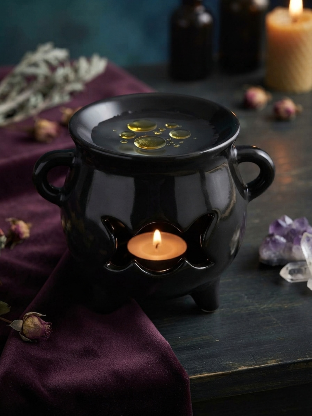A black cauldron-shaped candle wax warmer and oil burner with side handles and a lit candle inside, styled on a dark wooden surface with dried flowers, crystals, and other candles in the background.