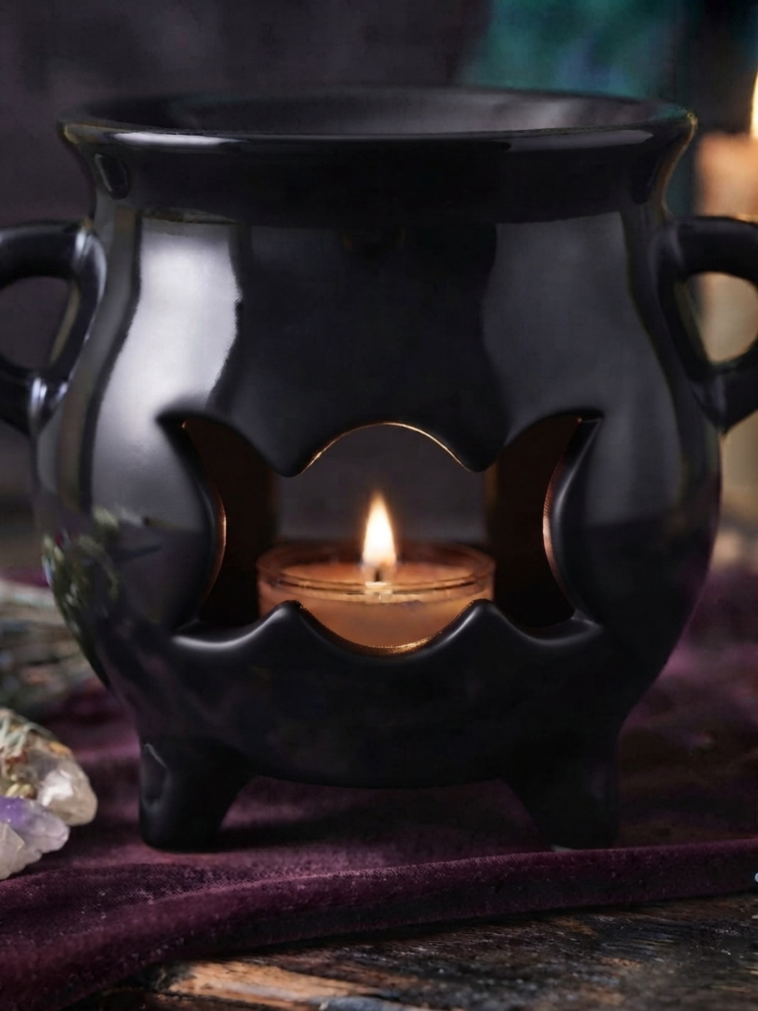 A glossy black cauldron-shaped wax warmer and oil burner with side handles and a triple moon cutout center, holding a lit tealight candle. The cauldron sits on a dark purple velvet cloth with a blurred mystical background.
