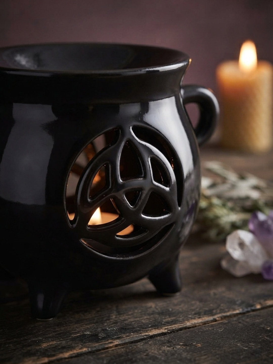 A black ceramic triple-triangle cauldron-shaped candle oil burner with a handle and tripod feet, shown lit on a rustic wooden surface with a blurred lit candle in the background.
