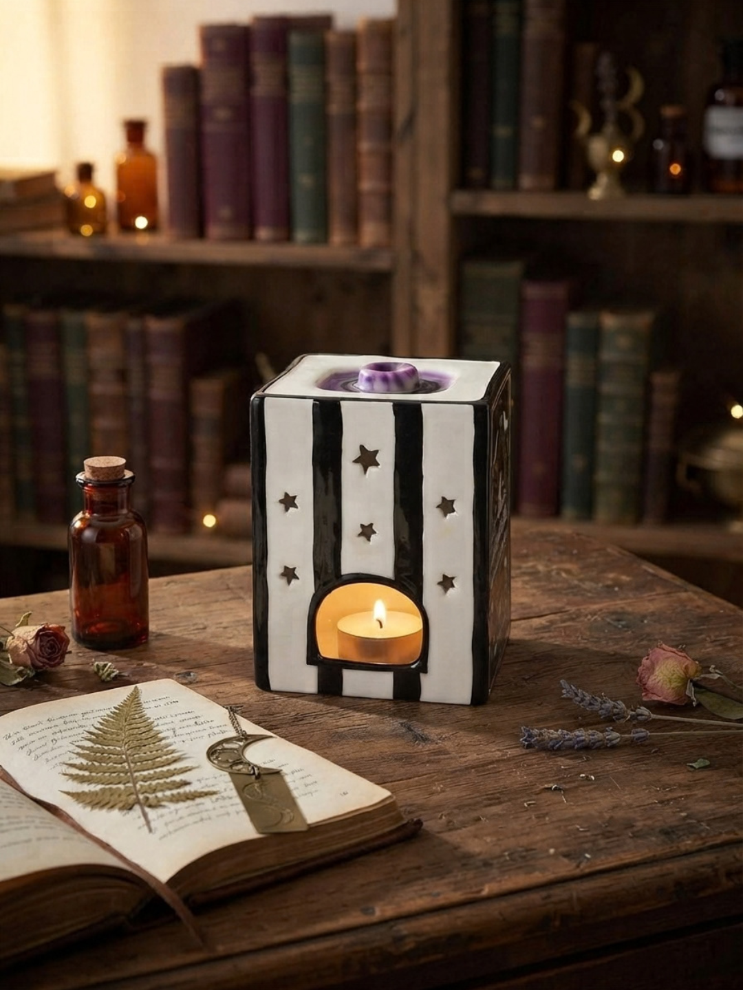 A black and white striped ceramic candle warmer with star cutouts and a glowing tealight, styled on a rustic wooden table next to an open book, dried flowers, and an amber glass bottle in a cozy library setting.